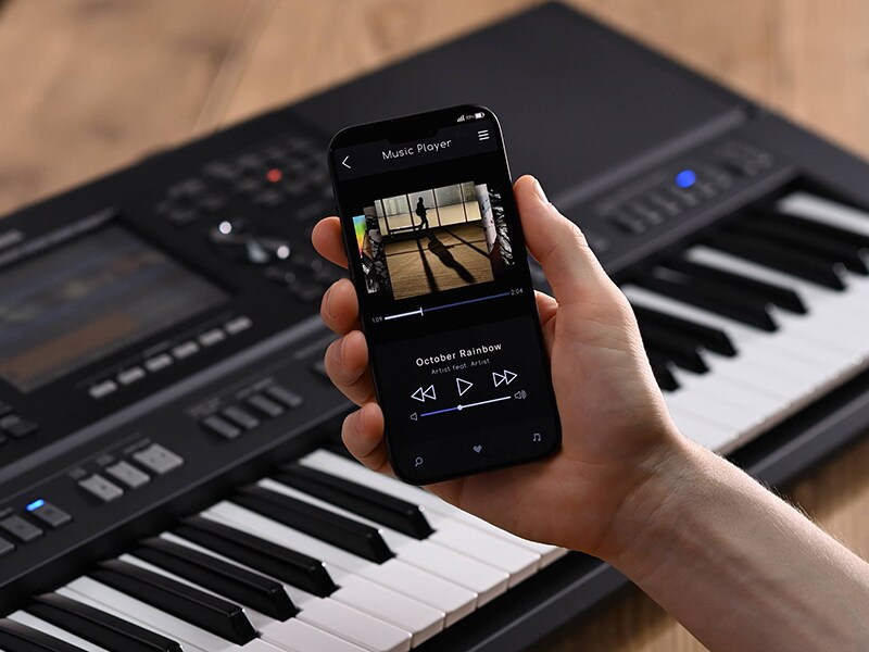 Yamaha Arranger Workstation PSR-SX920 og en hånd som holder en smarttelefon med en musikkspillerskjerm