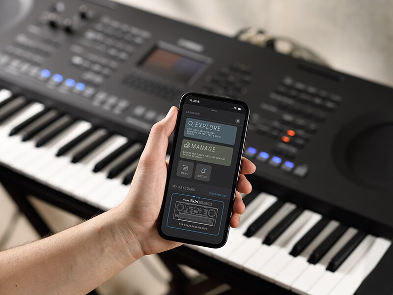 PSR-SX920 og en hånd som holder en smarttelefon med et musikkspillerskjermbilde.