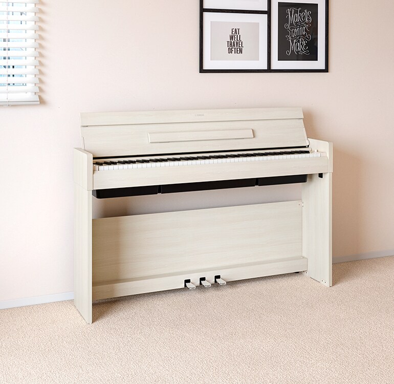 En stue med et ARIUS-piano i hvit bjørk plassert ved siden av vegghyller med lamper og en diffusor, harmonisk integrert i interiøret.